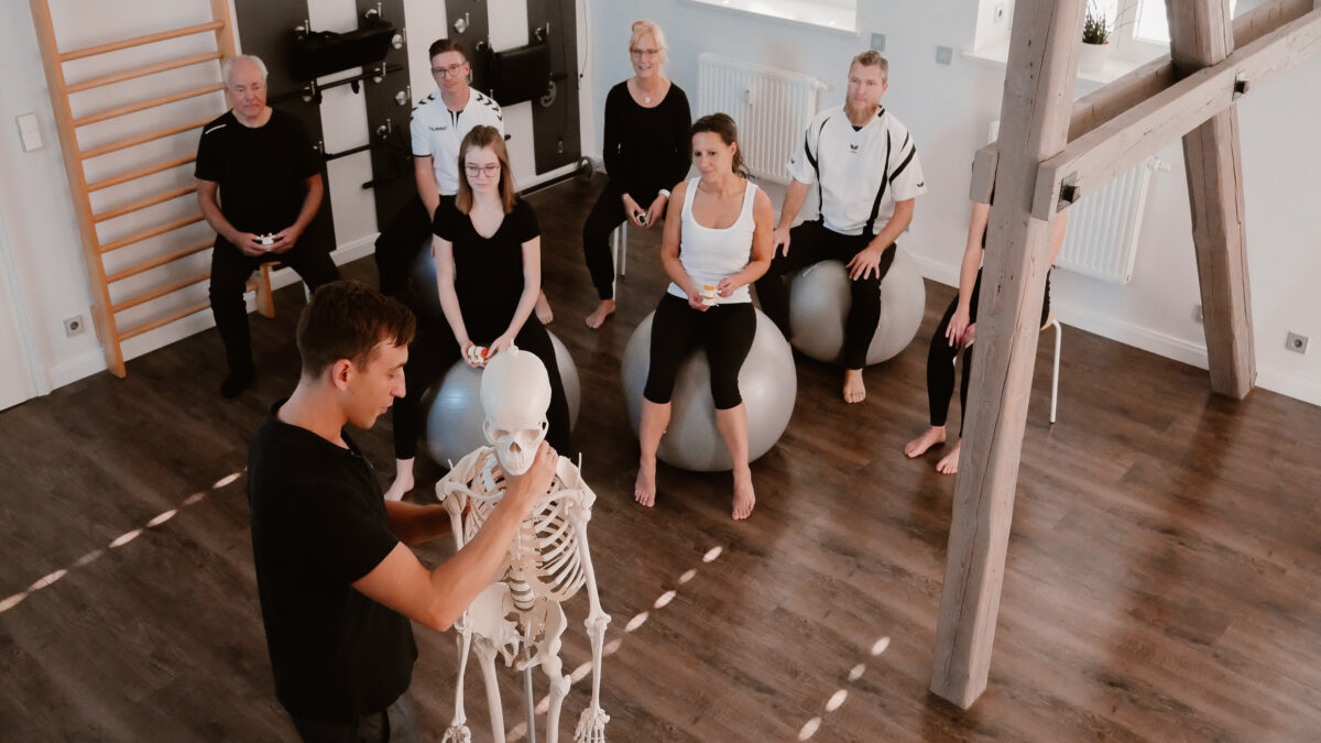Praeventionskurse Rückenschule: Kursleiter erklaert etwas am Demo-Skelett und Teilnehmer:innen schauen sitzend zu